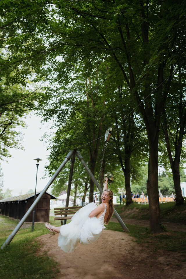 Braut auf Schaukel – verspielter Moment mitten im Grünen, Hochzeitsfoto mit Leichtigkeit