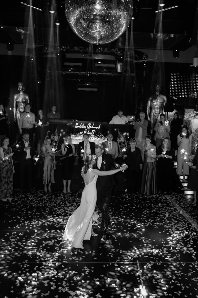 Hochzeitsfotografie von Patricia Bakker: First Dance unterm Discokugel-Licht, Gäste mit Wunderkerzen im Hintergrund (Schwarzweiß).