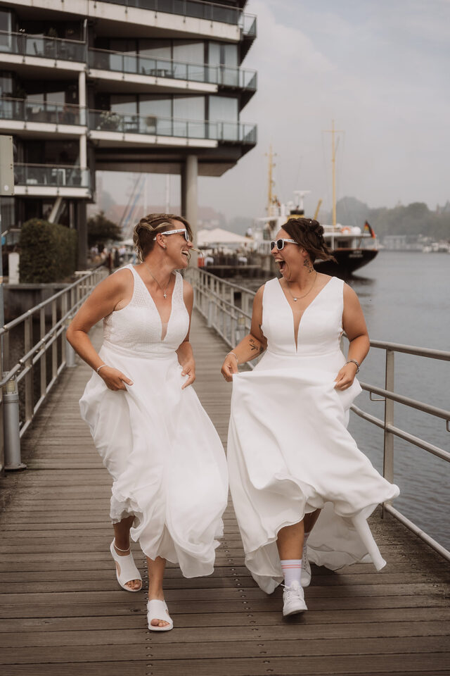 Lachend am Hafensteg: zwei queere Bräute im Fun-Moment – Hochzeitsreportage von Patricia Bakker (Hell Yes! Weddings).