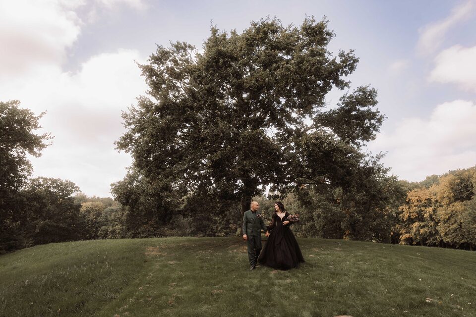 Weitwinkel-Ruhe: Gothic-Brautpaar spaziert über die Wiese unter großem Baum – Paarfoto von Patricia Bakker für natürliche Hochzeitsreportagen.