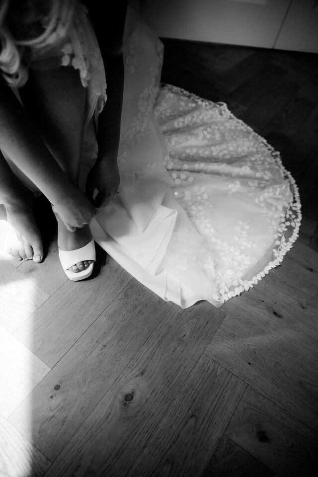 Der Moment vorm Losgehen: Braut zieht die Schuhe an, Kleidsaum im Licht – Hochzeitsreportage von Patricia Bakker (Schwarzweiß).