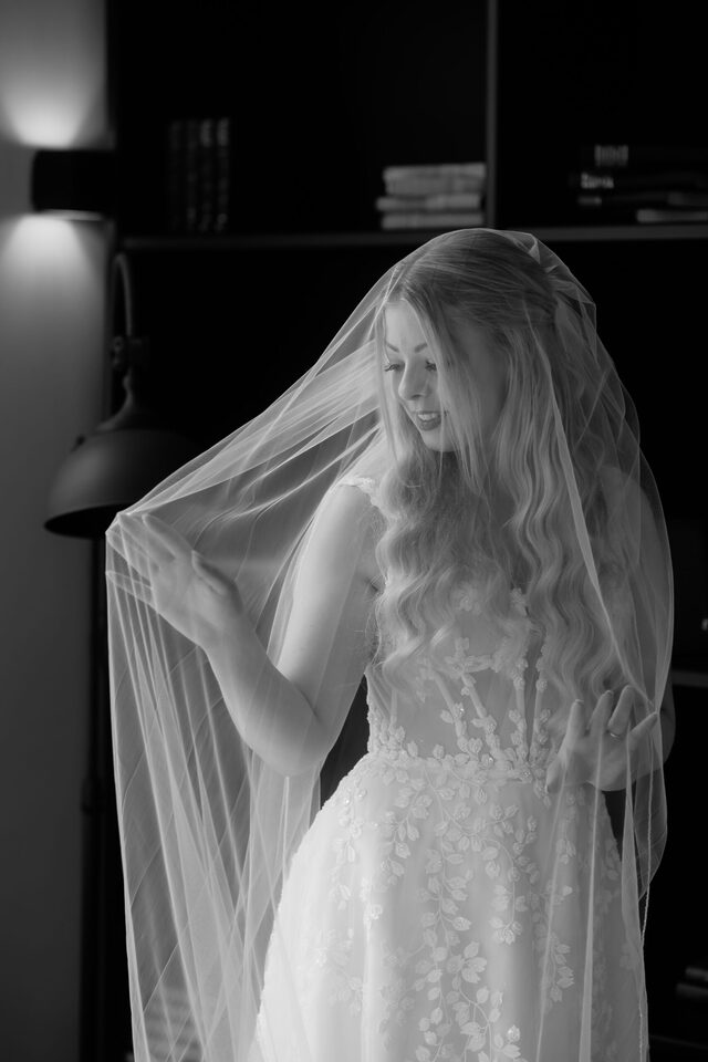 Ruhiges Brautportrait beim Getting Ready: Schleier-Spiel im Fensterlicht – Schwarzweißfoto von Patricia Bakker.