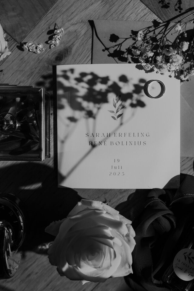 Hochzeitsfotografie von Patricia Bakker: Papeterie-Flatlay mit Einladung, Ringen und floralen Details – Hochzeit am 19. Juli 2025 (Schwarzweiß).