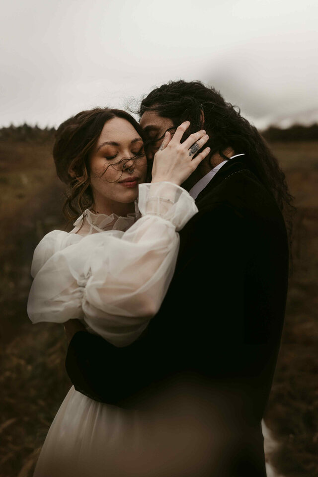 Ein Paar in inniger Umarmung in einer windigen Landschaft, beide mit geschlossenen Augen und vom Wind zerzaustem Haar, aufgenommen bei ihrem Elopement vom Elopement- und Hochzeitsfotografen Sturmsucht Dieses Farbbild, aufgenommen von "Elopement- und Hochzeitsfotograf Sturmsucht", zeigt ein Paar in einer innigen Umarmung während ihrer Elopement-Hochzeit in einer weiten, offenen Landschaft unter einem bewölkten Himmel. Die Frau, links im Bild, hat welliges braunes Haar, das teilweise vom Wind um ihr Gesicht weht, und ihre Augen sind geschlossen. Sie trägt ein helles, möglicherweise weißes oder cremefarbenes Kleid mit voluminösen, durchsichtigen Ärmeln und einem rüschenbesetzten Halsausschnitt. Sie hat einen Nasenring und leichte Sommersprossen sind auf ihrer Haut sichtbar. Ihr Arm ist um den Mann gelegt, und ihre Hand berührt sanft seinen Kopf. Der Mann, rechts im Bild, hat langes, dunkles, gelocktes Haar, das ebenfalls vom Wind bewegt wird. Er trägt einen dunklen Anzug oder Mantel über einem hellen Hemd, dessen Kragen sichtbar ist. Seine Augen sind ebenfalls geschlossen, und er hält die Frau fest in seinen Armen. An seinem Finger ist ein Ring mit einem detaillierten Muster zu sehen. Der Hintergrund ist eine weite, offene Fläche mit niedriger Vegetation in Brauntönen, die in der Ferne in einen dunklen Wald übergeht. Der Himmel ist hell und bewölkt, was der Szene eine weiche, diffuse Beleuchtung verleiht und die emotionale Verbundenheit des Paares hervorhebt.