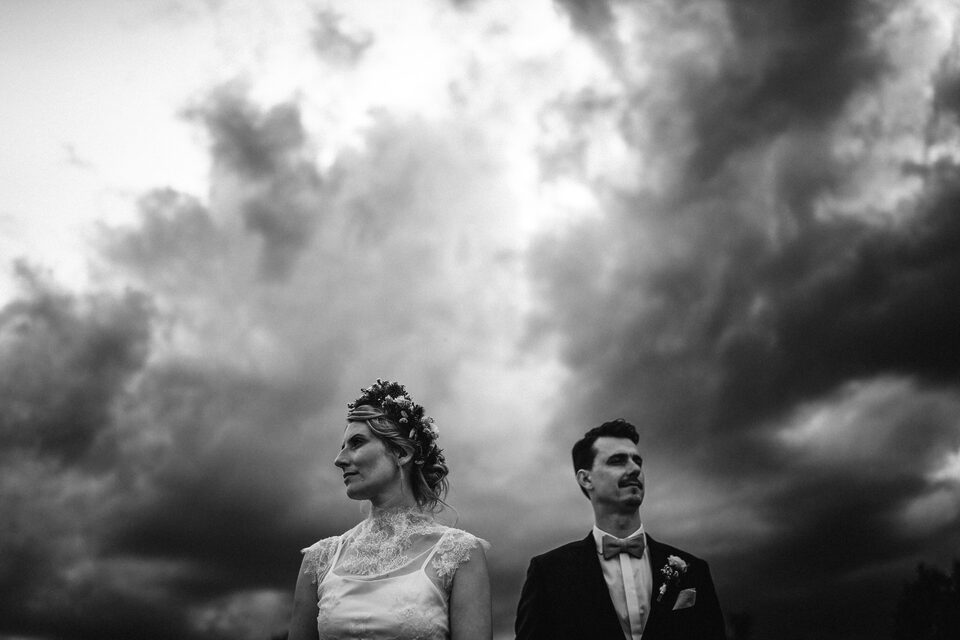 Ein Paar bei ihrem Elopement mit Hochzeitsfotograf Sturmsucht, die Braut in einem Spitzenkleid mit Blumenkrone und der Bräutigam im Anzug, die vor einem dramatisch bewölkten Himmel in entgegengesetzte Richtungen blicken. Dieses Schwarz-Weiß-Bild, aufgenommen vom Elopement- und Hochzeitsfotograf Sturmsucht, zeigt ein Elopement-Paar, das vor einem dramatischen, bewölkten Himmel steht. Die Braut, links im Bild, trägt ein helles, spitzenbesetztes Kleid und eine Blumenkrone im Haar, während sie nach links blickt. Der Bräutigam, rechts im Bild, trägt einen dunklen Anzug mit Fliege und einer Ansteckblume und blickt nach rechts oben, wodurch eine nachdenkliche oder abwartende Stimmung entsteht.