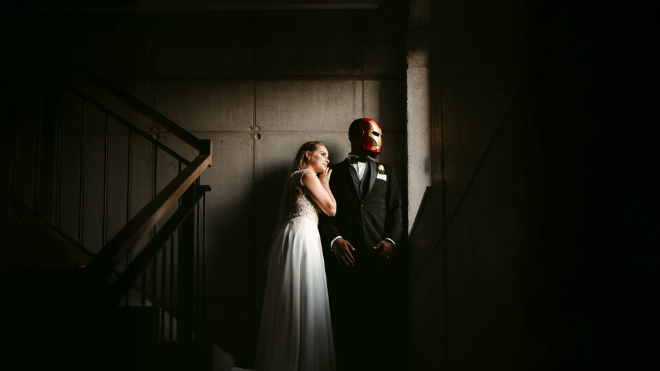 Ein Paar, fotografiert vom Elopement- und Hochzeitsfotograf Sturmsucht, steht während ihres Elopements in einem Raum mit Betonwänden und einer Treppe; die Braut trägt ein helles Kleid und blickt den Bräutigam an, der einen Smoking und eine rote Iron Man-Maske trägt. Dieses Farbbild, aufgenommen vom Elopement- und Hochzeitsfotograf Sturmsucht, zeigt ein Elopement-Paar in einem Innenraum mit Betonwänden und einer Treppe, wobei die Braut in einem hellen Kleid mit Spitzenapplikationen den Bräutigam ansieht, der einen eleganten Smoking trägt, aber anstelle seines Gesichts eine leuchtend rote und goldene Iron Man-Maske aufhat und zur Seite blickt.