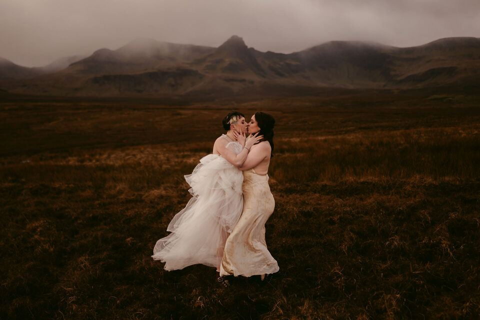 Queeres Elopement in Schottland: Kuss vor Berglandschaft aufgenommen von Hochzeitsfotograf Sturmsucht. Dieses Farbbild zeigt ein queeres Paar, zwei Frauen, die sich während ihres Elopements mit Hochzeitsfotograf Sturmsucht innig in einer weiten, rauen schottischen Landschaft küssen. Eine Frau trägt ein voluminöses, helles Hochzeitskleid, die andere ein enganliegendes, hellbraunes Kleid, vor einem Hintergrund aus nebligen Bergen und dunkler, karger Vegetation.
