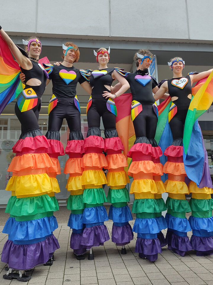 Fünf Stelzenläufer:innen mit bunten Regenbogenkostümen