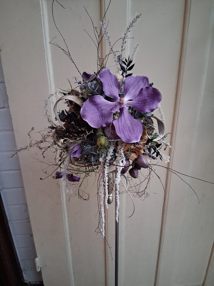 Skulpturaler Blumenstrauß mit großer lilafarbener Orchidee, dunklen Blättern, Zweigen und hängenden Details