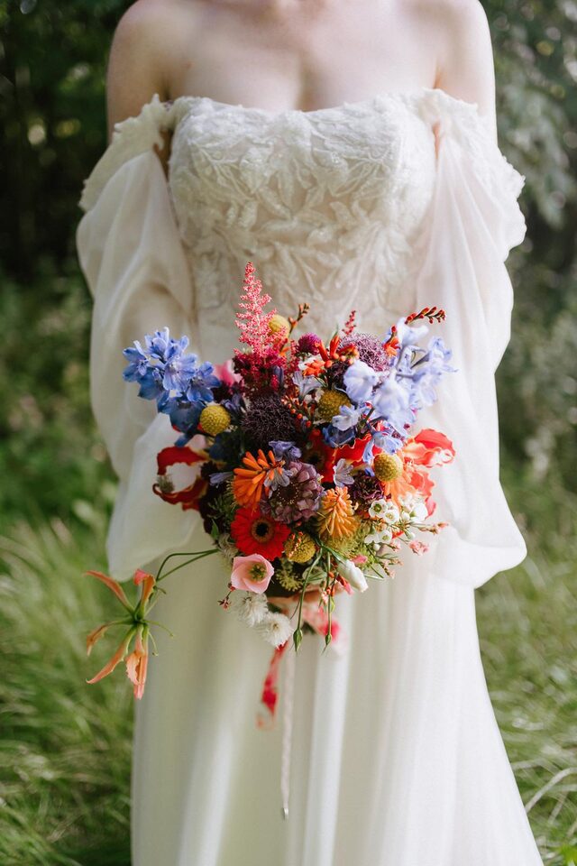 Person im weißen Hochzeitskleid mit locker gebundenem Brautstrauß in Blau, Orange, Pink und Gelb