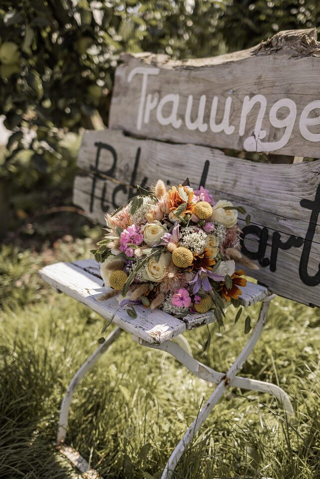 Bunter Brautstrauß in Rosa, Orange, Gelb und Creme vor einem rustikalen Holzschild zur Trauung