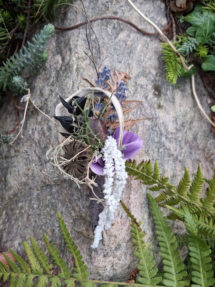 Florales Kunstwerk in Lila, Schwarz und Weiß mit Naturmaterialien auf einem Stein zwischen grünen Farnen
