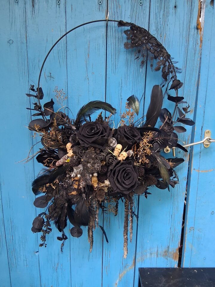 Runder Gothic-Blumenkranz mit schwarzen Rosen, Federn, Spitze und kleinen Totenköpfen an einer blauen Holztür