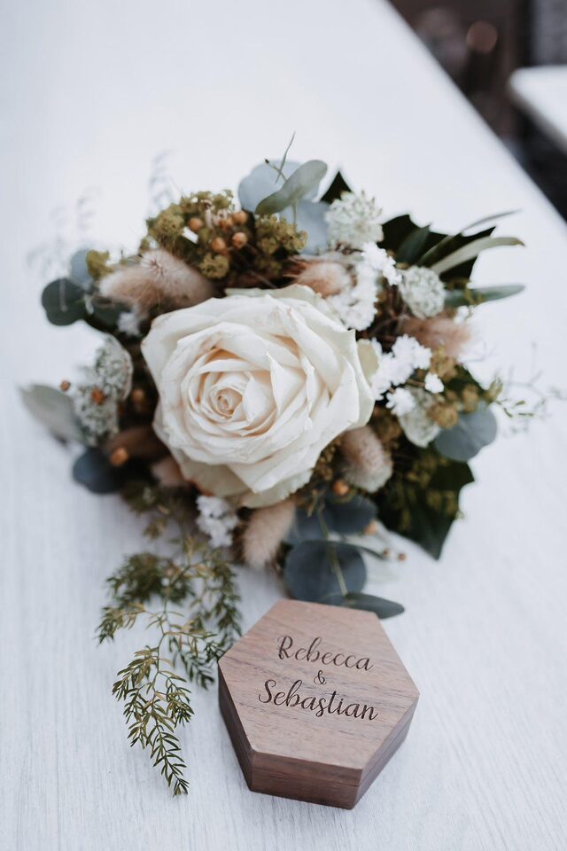 Weißer Brautstrauß mit Rose, Eukalyptus und Gräsern neben einer sechseckigen Ringschatulle aus Holz