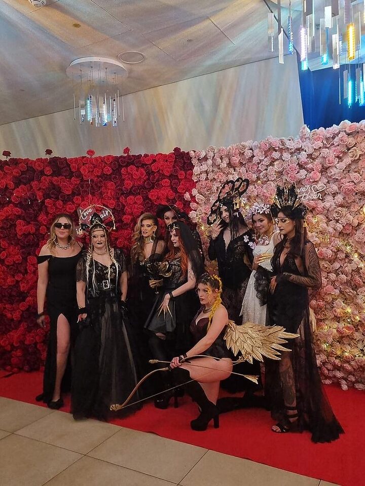 Gruppe in dunklen Outfits mit auffälligem Kopfschmuck vor einer rosa-roten Blumenwand auf einem roten Teppich