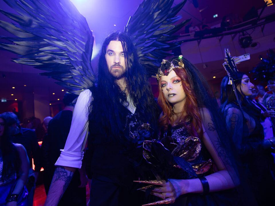 Zwei Personen in dunklen Fantasy-Outfits mit Federflügeln und Krone bei blau-violettem Veranstaltungslicht