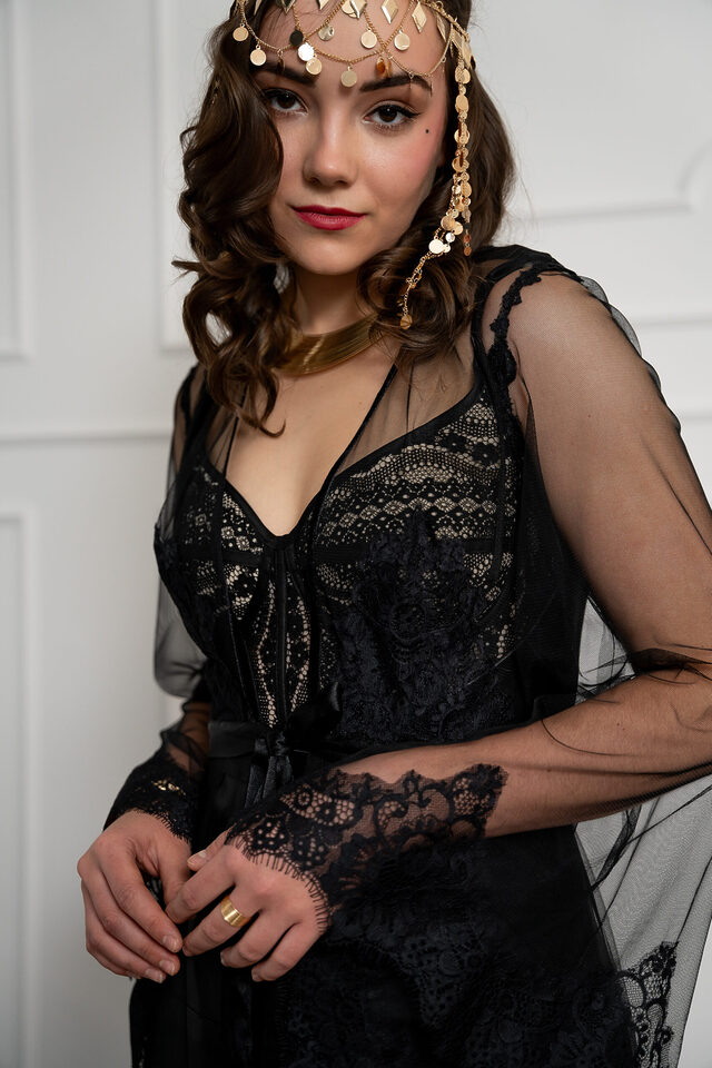 Person in schwarzem Spitzenoutfit mit transparenten Ärmeln und goldfarbenem Stirnschmuck
