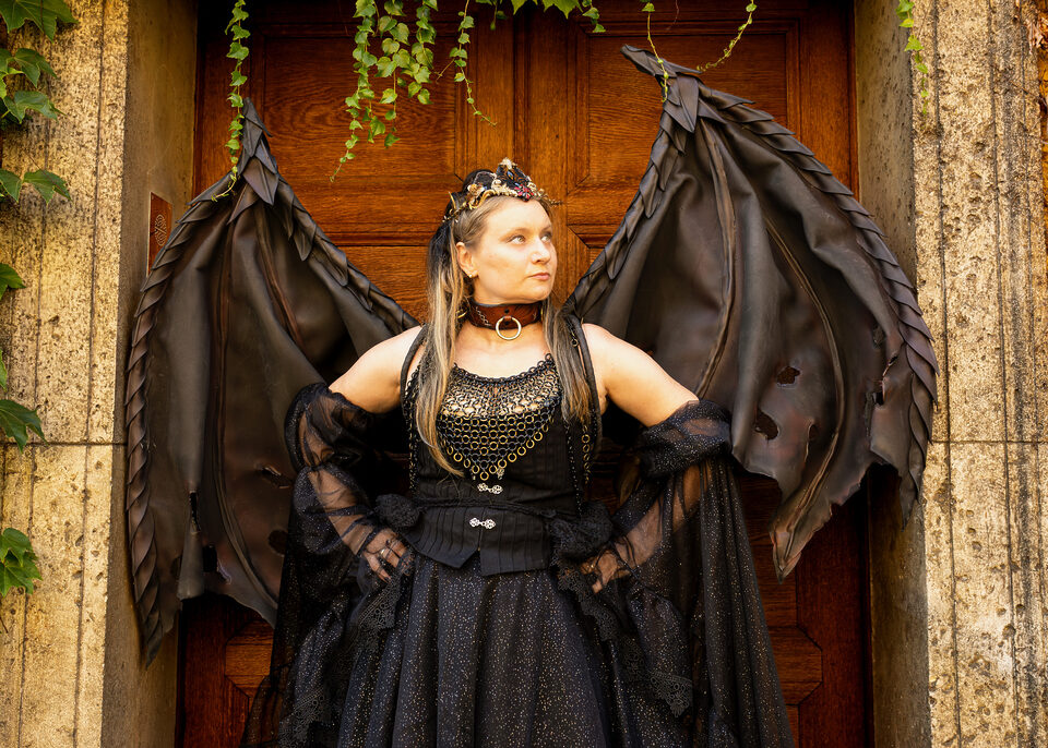 Person in schwarzem Fantasy-Outfit mit Krone, goldfarbenem Kettenschmuck und großen Drachenflügeln vor einer Holztür