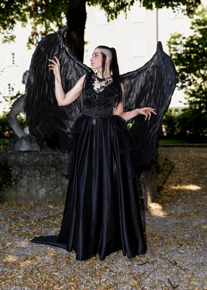 Person in langem schwarzem Kleid mit großen schwarzen Federflügeln unter einem Baum