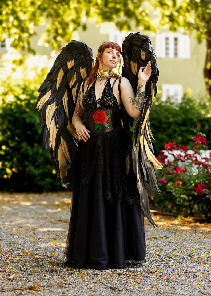 Person im schwarzen Outfit mit schwarz-goldenen Federflügeln und einer roten Rose an der Taille
