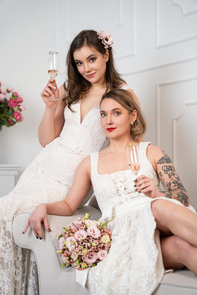 Zwei Personen in weißen Hochzeitsoutfits mit Sektgläsern, eine sitzend mit Blumenstrauß