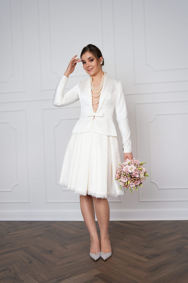 Person in weißem Blazer und kurzem weißem Rock mit Perlenketten und Blumenstrauß