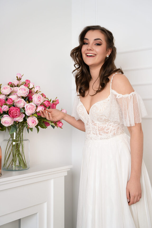 Person im weißen Brautkleid mit transparenten Schulterärmeln neben rosa Rosen in einer Glasvase