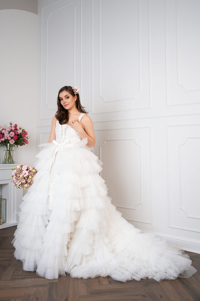Person in weißem Brautkleid mit voluminösem Stufenrock und langer Schleppe vor einer weißen Wand