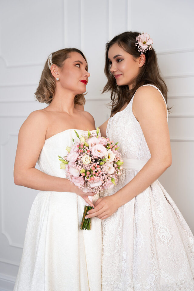 Zwei Personen in weißen Hochzeitsoutfits halten gemeinsam einen rosa-weißen Blumenstrauß