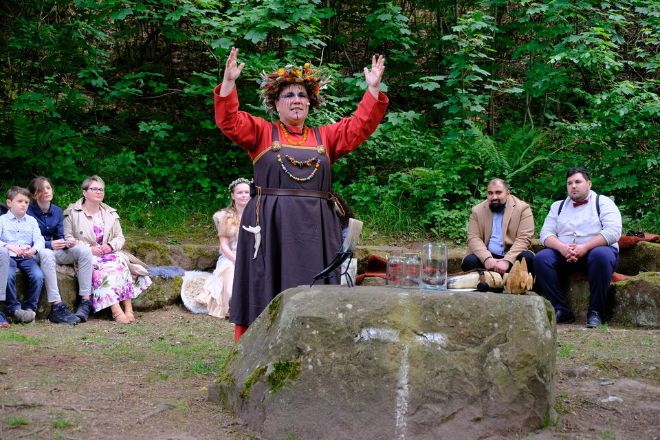 Freie Rednerin Nicole steht mit weit erhobenen Armen vor der Hochzeitsgesellschaft auf einem von Wald umgebenen Platz. Vor ihr befindet sich ein großer moosbewachsener Felsen mit Gefäßen für die Zeremonie. Brautpaar und Gäste sitzen im Halbkreis um sie herum und verfolgen die Wikingerhochzeit.