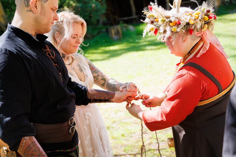 Nicole führt bei einer alternativen Hochzeit ein Handfasting-Ritual durch und bindet ein geflochtenes Band um die miteinander verbundenen Hände des Hochzeitspaares. Das tätowierte Paar trägt schwarz-weiße, historisch inspirierte Hochzeitskleidung und steht mit Nicole auf einer sonnigen Wiese.