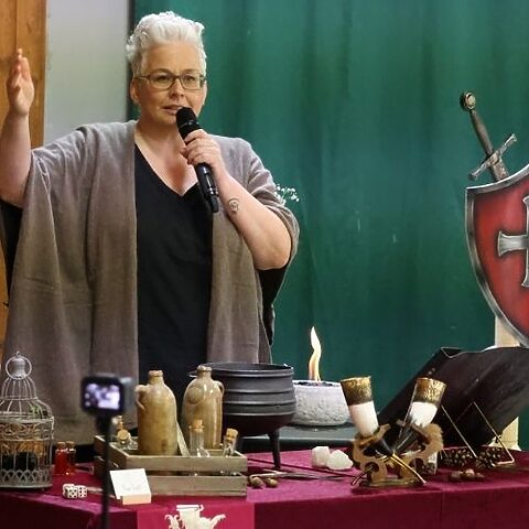 Tanja Rauth spricht mit Mikrofon vor einer mittelalterlich gestalteten Kulisse mit Schild, Schwert, Kerzen und Kessel.