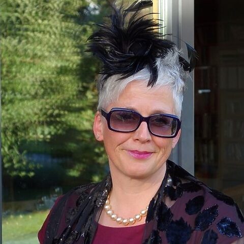 Porträt von Tanja Rauth mit kurzer weißer Frisur, Sonnenbrille und schwarzem Fascinator.