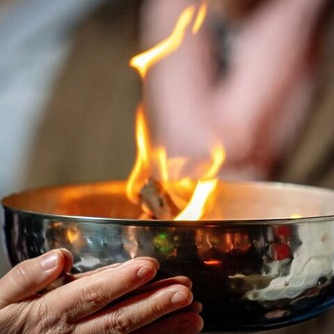 Nahaufnahme einer Hand, die eine silberne Schale mit brennender Flamme hält.